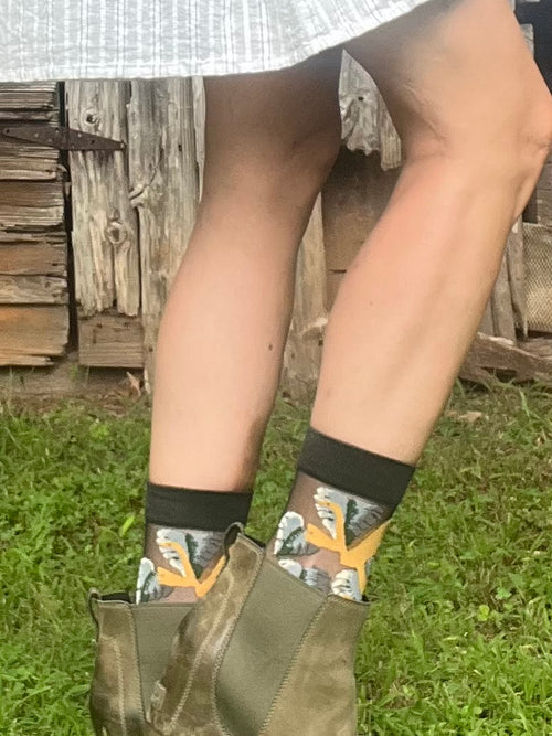 A person outdoors wearing patterned crew socks with a black cuff, paired with olive green boots and a gray skirt, standing on grass in front of a rustic wooden fence.