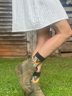 A person outdoors wearing olive green suede ankle boots and patterned socks, with a gray skirt visible above. The background shows grass and a rustic wooden wall.